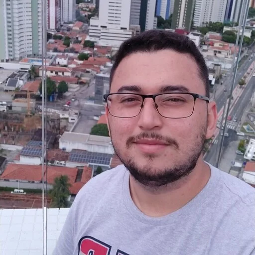 Portrait of Vinicius Gomes wearing glasses, in a grey t-shirt, with a city skyline behind him